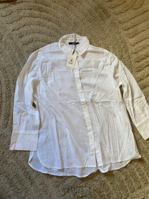Vit linneskjorta från Bik Bok - Klassisk vit skjorta från Bik Bok i luftigt linne. Skjortan har lång ärm, knappar framtill och en avslappnad passform. Perfekt till jeans eller kjol för en clean och fräsch look.