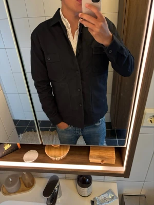 Svart overshirt från Zara - Säljer en svart overshirt från Zara i relaxed fit. Skjortjackan har två bröstfickor med lock och knappar samt två sidofickor. Perfekt att bära över en t-shirt eller skjorta för en avslappnad men stilren look.