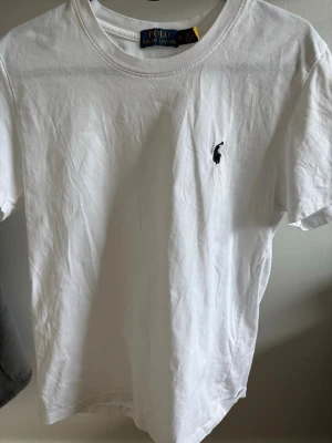 Vit t-shirt från Polo Ralph Lauren - Klassisk vit t-shirt från Polo Ralph Lauren med rund halsringning och ikonisk broderad logga på bröstet. Tillverkad i mjuk bomull och har en normal passform. Perfekt basplagg för en stilren look.