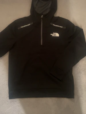 Svart hoodie från The North Face - Säljer en svart hoodie från The North Face med halv dragkedja och vit logga på bröstet. Hoodien har huva och detaljer med The North Face-tryck på axlarna. Tillverkad i mjukt och bekvämt material, perfekt för chill dagar eller när du vill ha en clean look.