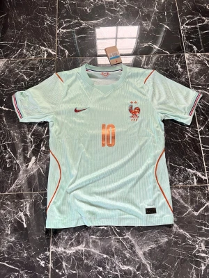 Ljusblå Frankrike Nike matchtröja - Snygg ljusblå matchtröja från Nike med Frankrikes landslagsemblem och nummer 10 i orange framtill. Tröjan har korta ärmar, rund hals och tunna ränder i tyget. Orange och röda detaljer vid axlar och ärmslut. Perfekt för fotbollsfans!