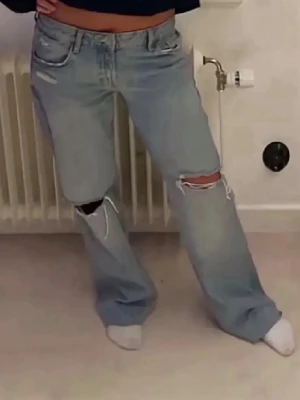 Ljusa slitna raka jeans från Zara - Säljer ett par ljusblå raka jeans från Zara med slitningar och hål på båda knäna. Jeansen har fem fickor, hög midja och klassisk dragkedja med knapp. Perfekta för en avslappnad och trendig look.
