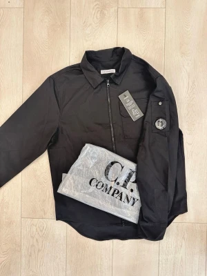 Svart overshirt från C.P. Company - Stilren svart overshirt från C.P. Company med klassisk krage, dragkedja framtill och en ficka på bröstet. Ärmen har den ikoniska linsen på armen. Tillverkad i ett lätt och slitstarkt material, perfekt för lager-på-lager och streetwear-vibe.