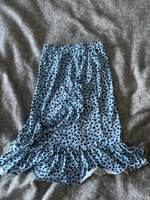 Blå prickig volangkjol - Supersöt blå kjol med svarta prickar och volangkant. Kjolen har en omlott-look med volangdetalj framtill och resår i midjan för skön passform. Perfekt för dig som gillar lekfulla mönster och trendiga detaljer.