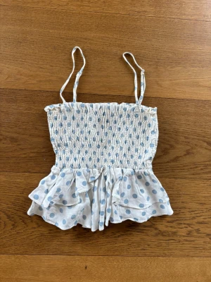 Prickig volangtop från Zara - Supersöt topp från Zara med ljusblå prickar på vit botten. Smockad överdel, tunna axelband och volang nedtill ger en lekfull vibe. Perfekt för varma dagar och sommarens alla äventyr.