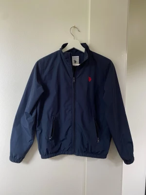 Marinblå vindjacka U.S POLO ASSN - Snygg mörkblå vindjacka från U.S POLO ASSN. röd broderad logga på bröstet. Jackan har dragkedja framtill, två fickor med dragkedja och elastiska muddar vid ärmar och midja. Klassisk rak passform och hög krage. Perfekt för blåsiga dagar. Passar folk mellan 12-13 år.