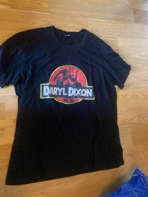 Svart Daryl Dixon t-shirt - Cool svart t-shirt med stort Daryl Dixon-tryck på bröstet i rött, gult och vitt. Motiv inspirerat av Jurassic Park-loggan. Klassisk rund hals och korta ärmar. Perfekt för fans av serien och streetwear.