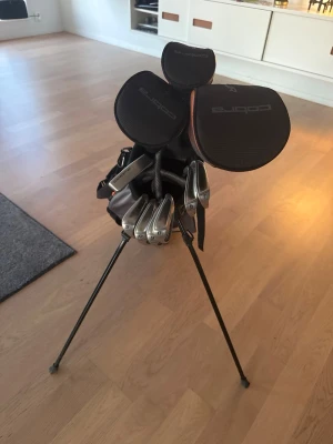 Golfset Cobra med bag och klubbor - Säljer ett komplett golfset från Cobra junior set 13-15 år med flera järnklubbor och tillhörande svart och grå golfbag med orange detaljer. Bagen har praktiska fack och benstativ för enkel hantering på banan. Perfekt för dig som vill komma igång med golf eller uppgradera din utrustning.