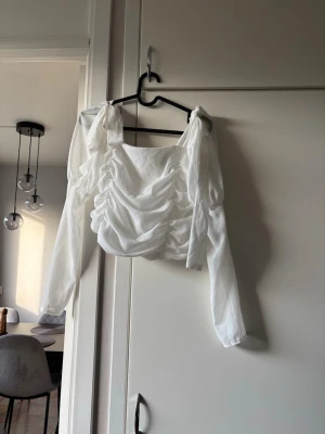 Vit omlottopp med dragkedja - Trendig vit topp med långa ärmar och snygg omlottdesign. Toppen har rynkade detaljer framtill och dragkedja i ryggen. Tillverkad i ett lätt, skirt material som ger en luftig känsla. Perfekt för dig som vill sticka ut med en unik och stilren look.