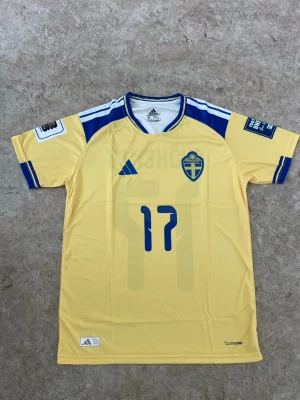 Svensk landslagströja Adidas nr 17 - Gul svensk landslagströja från Adidas med blå detaljer, nummer 17 och namnet Gyökeres på ryggen. Tröjan har korta ärmar, blått tryck och Climacool-material för bra ventilation. Perfekt för fotbollsfans som vill stötta Sverige.