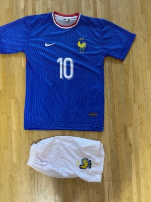 Frankrike fotbollströja Mbappé #10 - Säljer en blå fotbollströja från Nike med Frankrikes landslagsmärke och nummer 10, Mbappé på ryggen. Tröjan har vita detaljer och röd insida vid kragen. Tillhörande vita shorts med landslagslogga ingår. Perfekt för dig som älskar fotboll och vill stötta Les Bleus.