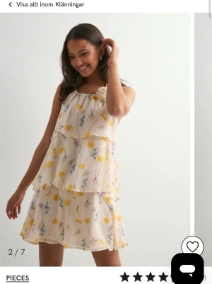 Blommig volangklänning från Pieces - Söt kort volangklänning från Pieces i ljusbeige med blommigt mönster i gult och blått. Klänningen är ärmlös, har lager på lager-effekt och en dekorativ blomdetalj vid halsen. Perfekt för sommarens alla tillfällen. Prislappen kvar 