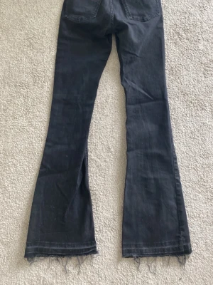 Svarta bootcut jeans med rå kant - Säljer ett par svarta bootcut jeans med rå, fransig kant nertill. Jeansen har hög midja och klassiska bakfickor. Materialet är mjukt denim som ger en skön passform och en avslappnad look. Perfekta för dig som gillar en trendig och lite retro stil.