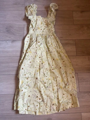 Ljusblommig långklänning med volang - Söt långklänning i ljusgul ton med blommigt mönster. Klänningen har volangdetaljer på axelbanden, knytning i ryggen och är ärmlös. Perfekt för sommarens varma dagar och har en romantisk känsla.