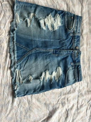 Blå jeanskjol från Hollister - Säljer en blå kort jeanskjol från Hollister med hög midja och slitna detaljer. Kjolen har fransig nederkant och flera revor framtill för en trendig, avslappnad look. Perfekt för dig som gillar streetstyle och denim.