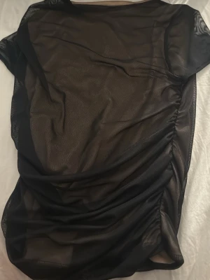 Svart mesh topp från ASOS DESIGN - Säljer en svart mesh topp från ASOS DESIGN. Toppen är genomskinlig, har korta ärmar och är lätt rynkad på ena sidan för en snygg effekt. Perfekt att styla lager på lager eller med ett linne under. Enkel och trendig design.