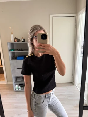 Svart topp med guldknappar från H&M - Svart croppad topp från H&M med korta ärmar och tre stora guldknappar på ena axeln. Materialet är strukturerat och känns lite tjockare, vilket ger toppen en lyxig känsla. Kom aldrig till användning.