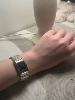 Silverfärgad klocka från Michael Kors - Stilren klocka från Michael Kors med rektangulär svart urtavla och silverfärgat metallarmband. Klassisk design med romerska siffror och diskret logga på urtavlan. Perfekt accessoar för dig som gillar en tidlös look.