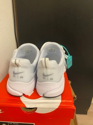 Nike Air Presto vita sneakers - Nike Air Presto sneakers i helvitt med klassisk swoosh-logga på sidan och AIR-tryck på hälen. Skorna har en sportig siluett, meshmaterial för bra andningsförmåga och vita snören. Sulan är flexibel med greppmönster och svart detalj undertill.