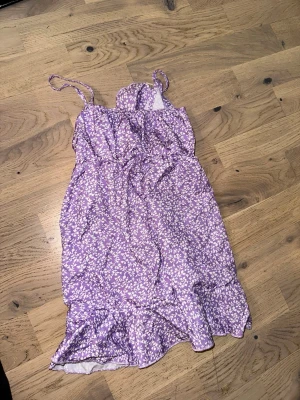 Lila blommig sommarklänning med volang - Lila blommig sommarklänning med volang Söt lila klänning med vita blommor, smala axelband och volang nertill. Perfekt för varma dagar och har en avslappnad passform. Tillverkad i ett lätt material som känns skönt mot huden.