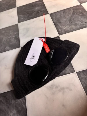 Svart mössa med goggles C.P. Company - Säljer en svart ribbstickad mössa från C.P. Company med ikoniska goggles framtill. Mössan har uppvikt kant och är tillverkad i mjukt, stretchigt material som sitter skönt på huvudet. Perfekt accessoar för dig som gillar streetwear och vill sticka ut.