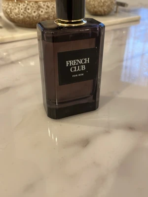 French Club For Him parfym - Stilren herrparfym från French Club i en mörklila, rektangulär glasflaska med svart etikett och gulddetaljer vid halsen. Flaskan har en modern och elegant design som passar perfekt på badrumshyllan.