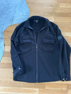 Svart overshirt från Cato - Stilren svart overshirt från Cato med dragkedja framtill och två stora bröstfickor med lock. Overshirten har en klassisk krage och knappdetaljer vid ärmsluten. Tillverkad i ett mjukt och värmande material, perfekt för lager på lager.