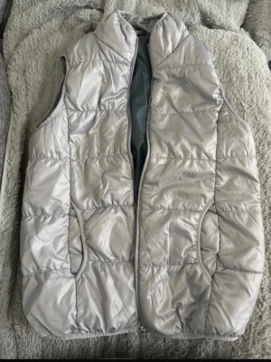 Ljusgrå quiltad dunväst - Snygg ljusgrå dunväst med quiltad design och hög krage. Västen har två fickor framtill och stängs med dragkedja. Materialet är glansigt och ger en modern look. Perfekt att slänga över en hoodie eller tröja för extra värme.