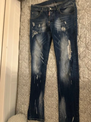 Blå  jeans - Säljer ett par blå skinny jeans med slitningar och hål på både fram- och baksida. Jeansen har klassisk femficksmodell, knappgylf och tydliga slitningar för en trendig look. Perfekta för dig som gillar en avslappnad och edgy stil.