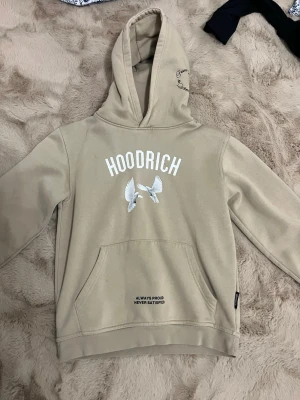 Beige hoodie från Hoodrich med tryck - Snygg beige hoodie från Hoodrich med vita duvor och texttryck både fram och bak. Stor huva, magficka och coola detaljer som 'ALWAYS PROUD NEVER SATISFIED' och 'From Nothing To Something'. Perfekt för dig som gillar streetwear och vill sticka ut.