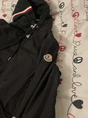 Svart Moncler vindjacka med huva - Snygg svart vindjacka från Moncler med dragkedja, huva och vita/röda detaljer på huvan. Jackan har Moncler-logga på ärmen och är tillverkad i ett lätt, vindtätt material. Perfekt för blåsiga dagar och enkel att matcha med olika outfits.