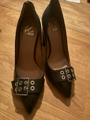 K.Cobler Svarta pumps, st.38 (24 cm) - Snygga svarta pumps från Cobbler med bred klack och spetsig tå. Skorna har ett dekorativt bältesspänne med metallnitar framtill. Tillverkade i läderimitation med en klassisk och edgy look.