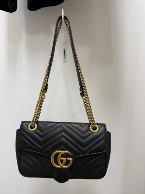 Gucci Marmot - Snygg svart handväska i quiltat läder med guldfärgade kedjor och GG-detalj framtill. Väskan har ett hjärtmönster på baksidan och justerbar axelrem i kedja och läder. Perfekt accessoar för att lyfta din outfit.
