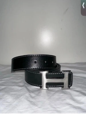 Svart bälte med silverfärgat spänne - Snyggt svart bälte i skinn med kontrasterande vita sömmar och ett stort, silverfärgat H-format spänne. Perfekt accessoar för att lyfta vilken outfit som helst. Klassisk design som funkar till många olika stilar.
