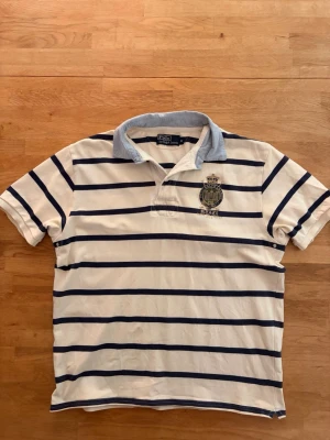Ralph Lauren Piké - Snygg vit pikétröja från Ralph Lauren med mörkblå horisontella ränder och ljusblå krage. Tröjan har korta ärmar och ett broderat emblem på bröstet. Tillverkad i bomull för en bekväm och klassisk look. Har sytts om vid midjan för att passa bättre. Skriv vid funderingar.