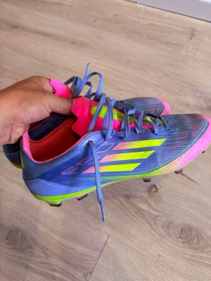 Blåa Adidas fotbollsskor neonrosa - Säljer ett par grymma Adidas fotbollsskor i blått med neonrosa och neongula detaljer. Skorna har snörning, låg profil och är tillverkade i syntetmaterial för bästa känsla på planen. Perfekta för dig som vill sticka ut på matchen!