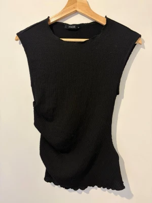 Svart ribbad ärmlös topp - Säljer en svart ribbad topp från Xside. Toppen är ärmlös med rund halsringning och har en stretchig, figurnära passform. Perfekt att styla med jeans eller kjol för en clean look.
