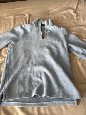 Ljusblå halvzip från Polo Ralph Lauren - Ljusblå ribbstickad tröja med halv dragkedja och klassisk Polo Ralph Lauren-logga broderad på bröstet. Tröjan har hög krage och långa ärmar, perfekt för lager på lager. Materialet ser ut att vara mjukt och bekvämt.