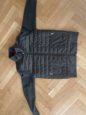 Hrön quiltad jacka från Iceland - Snygg mörkgrå quiltad jacka från Iceland med stickade ärmar och krage. Jackan har dragkedja framtill och två fickor med dragkedja. Insidan av kragen har en röd kant som ger en cool kontrast. Perfekt för kyliga dagar.
