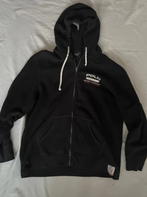 Vintage Polo Jeans Co. Ralph Lauren zip-up hoodie – svart - Svart hoodie från Polo Jeans Company med dragkedja framtill och vita dragsnören i huvan. Märket syns broderat på bröstet och en etikett nertill. Tillverkad i mjuk fleeceblandning av 80% bomull och 20% polyester. Praktiska kängurufickor framtill.