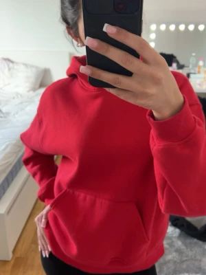 Röd hoodie med magficka - Säljer en klassisk röd hoodie med huva och stor magficka framtill. Hoodien har ribbade muddar vid ärmslut och nederkant, samt en avslappnad passform. Perfekt för dig som gillar en enkel och stilren look.