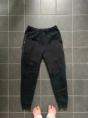 Svarta mjukisbyxor med dragkedja - Säljer ett par svarta mjukisbyxor med slim fit och ribbade muddar vid bensluten. Byxorna har en dragkedjeförsedd ficka på sidan och markerade sömmar för en sportig look. Perfekta för chill eller träning.