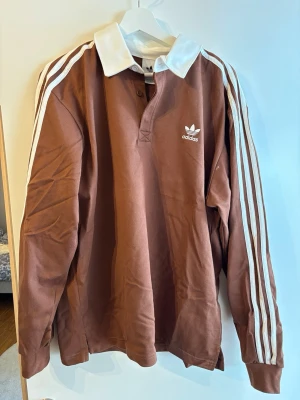 Brun långärmad tröja från Adidas - Säljer en brun långärmad tröja från Adidas med vit krage och klassiska vita ränder längs ärmarna. Tröjan har knappslå vid halsen och Adidas-logga på bröstet. Perfekt för en sportig och avslappnad stil.