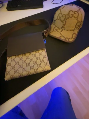 Beige handväska med GG-mönster - Snygg beige handväska med klassiskt GG-monogram i brunt. Väskan har en brun läderdetalj upptill och justerbar axelrem. Insidan har två fack och är fodrad i brunt tyg. Perfekt för dig som gillar lyxiga accessoarer och tidlös design.