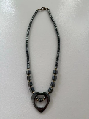 Halsband med hjärthänge och pärlor - Snyggt halsband i metallisk grå färg med ett hjärthänge och små pärlor i mitten. Halsbandet består av cylinderformade pärlor och kubformade detaljer som ger ett coolt och modernt intryck. Perfekt accessoar för att lyfta din outfit.