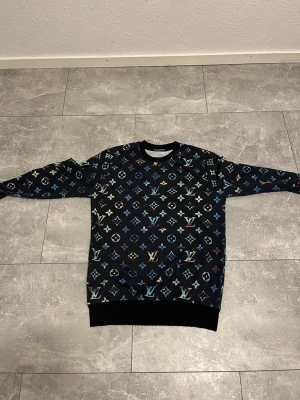 Louis Vuitton svart sweatshirt L - Säljer en svart sweatshirt från Louis Vuitton i storlek L. Tröjan har ett färgglatt all-over monogrammönster i blått, grönt, gult och rosa. Rund halsringning och ribbade muddar vid ärmslut och nederkant. Perfekt för dig som gillar streetwear och exklusiva märken.