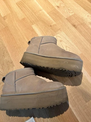 Beiga plattformstövlar från UGG - Säljer ett par beiga UGG-stövlar med plattformsula och mjukt foder. Skorna är i mocka och har en låg, rundad siluett med dragflik baktill. Perfekta för att hålla värmen och ge extra höjd till din look. Vattenskador och lite smutsiga på sulan annars ingen slitage.