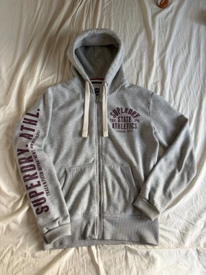 Grå zip hoodie från Superdry - Snygg grå zip hoodie från Superdry med tryck på bröstet och längs ena ärmen. Hoodien har vita dragsnören, två fickor fram och mjukt fleecefoder på insidan. Perfekt för en avslappnad och sportig stil.