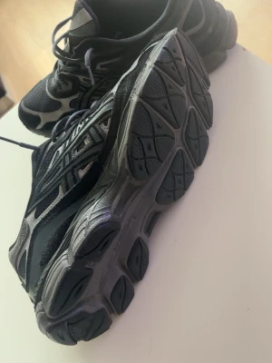 Svarta och grå Asics sneakers - Snygga sneakers från Asics i svart och grått med meshpaneler och detaljer i syntet. Skorna har en chunky sula och klassisk snörning. Perfekta för dig som gillar sportig stil och vill ha bekväma skor till vardags.