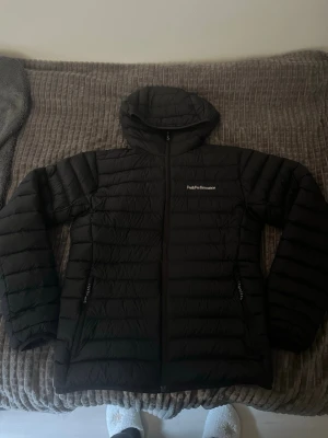 Svart pufferjacka från Peak Performance - Snygg svart pufferjacka från Peak Performance med huva och dragkedja framtill. Jackan har två sidofickor med dragkedja och diskret logga på bröstet. Perfekt för kalla dagar och riktigt skön att bära. Klassisk quiltad design och lätt vaddering.